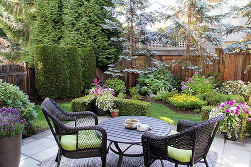 How to enjoy fall in the backyard.  Image of a backyard with table, two chairs big green plants and flowers. 